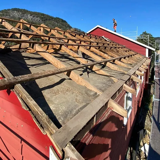 Bart tak i forgrunnen, med arbeider i bakgrunnen oppe på et annet, høyere tak. Blå himmel, berg og sjø kan skimtes i bakgrunnen også.