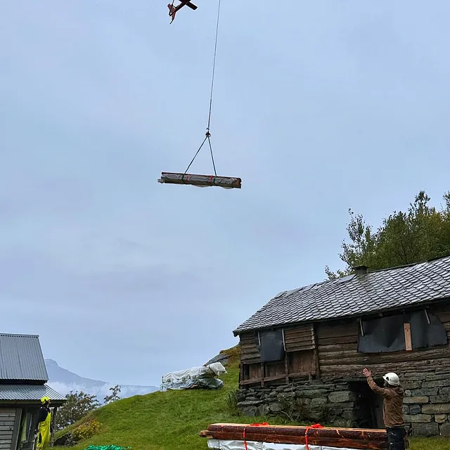 Et helikopter frakter bygningsmaterialer hengt i en line. To personer på bakken gir veiledning ved gamle trehus i et grønt landskap.