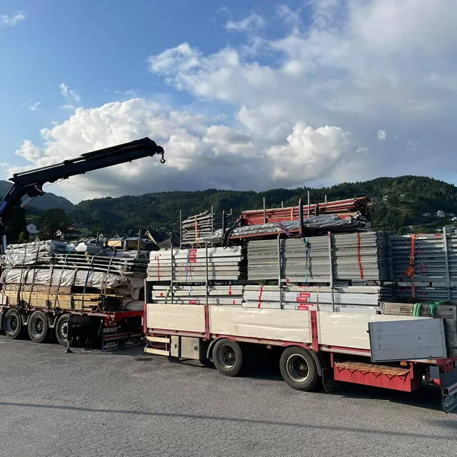 En lastebil med kran laster bygningsmaterialer. Materialene er sikret på tilhengeren, og i bakgrunnen er det fjell og spredte hus.